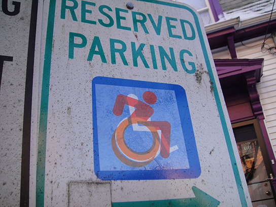 This header photo shows the earliest street-art version of the icon: a clear-backed sticker with an orange and red wheelchair-riding icon, superimposed over a blue and white standard icon. Old and new together.